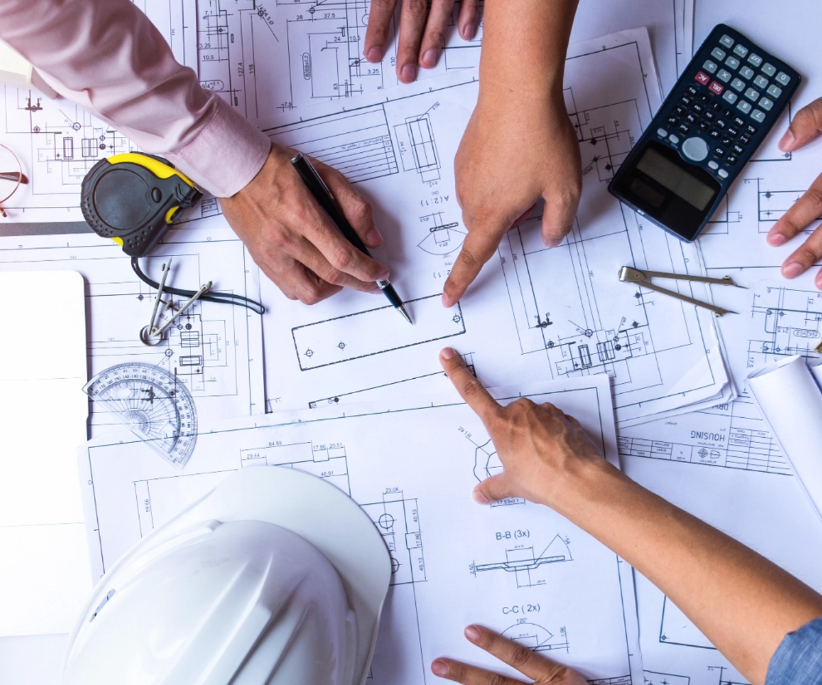 Hands pointing at architectural plans and blueprints, with tools and a calculator on a table.